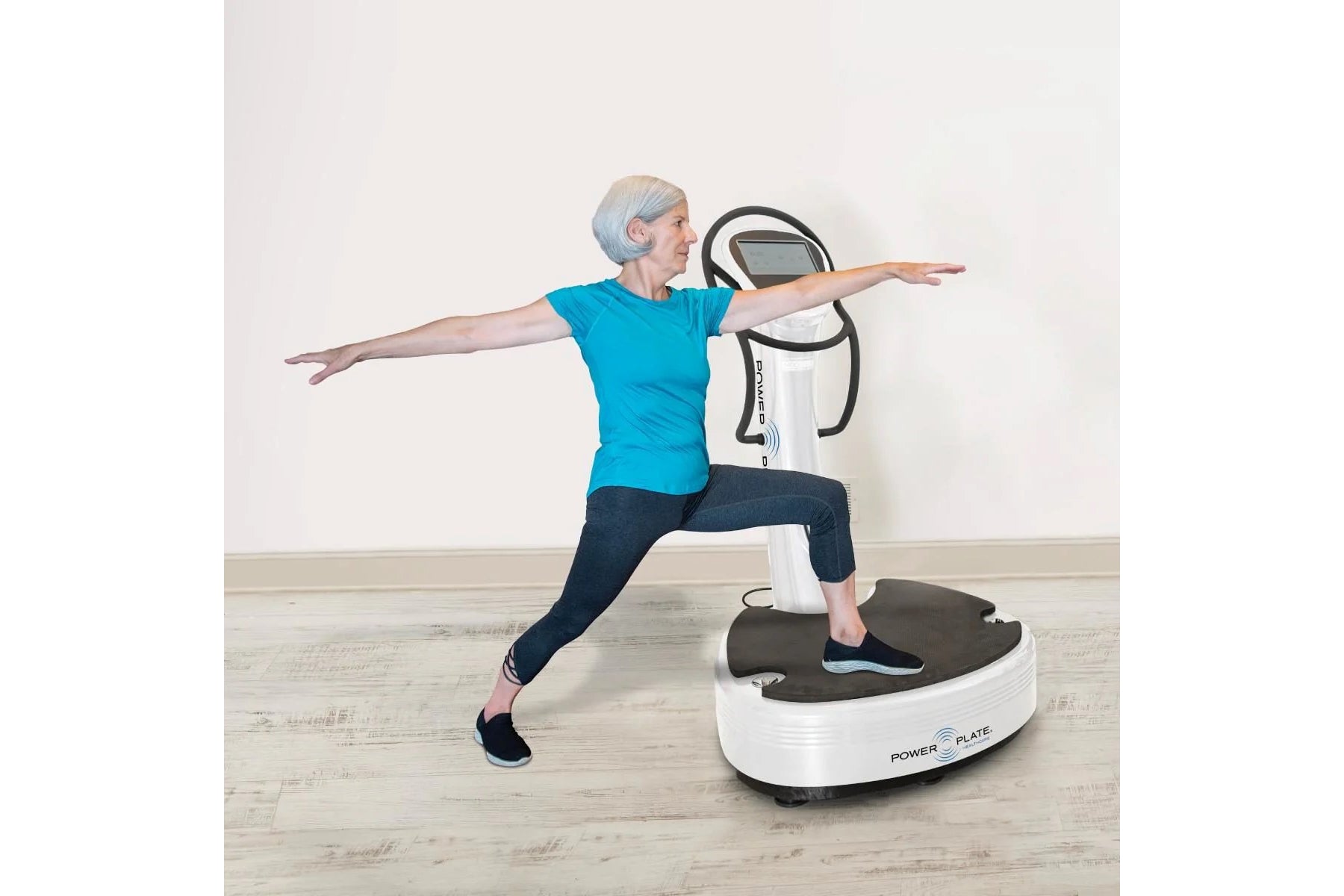 Senior woman in bright blue t-shirt and navy leggings exercising on white Power Plate vibration trainer with arms extended in