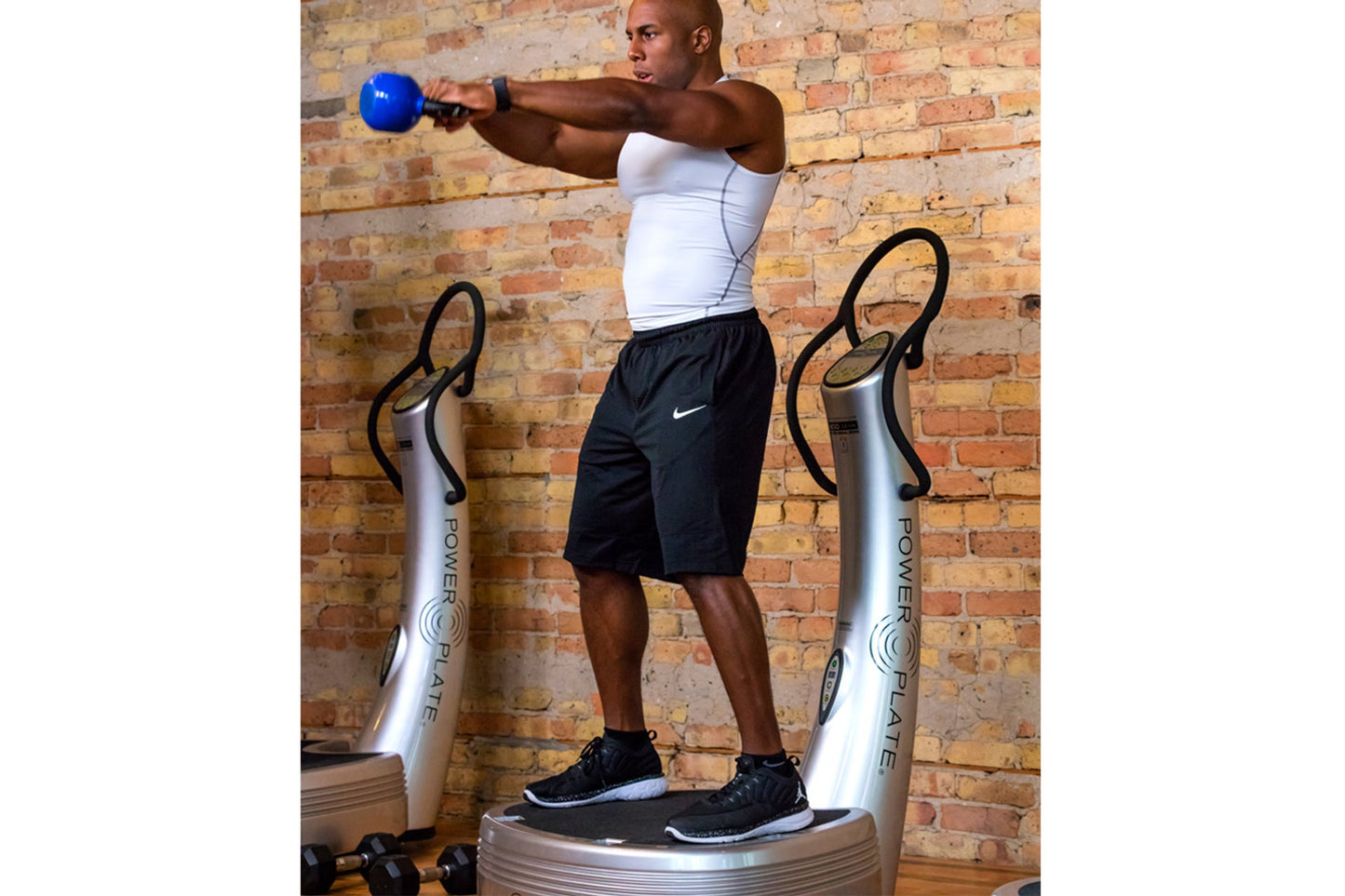 Man using silver Power Plate vibration trainer while holding blue kettlebell, wearing white tank top and black shorts against
