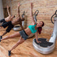 Woman in turquoise sports bra and gray shorts performing side plank on vibration plate machine in gym studio with brick wall 
