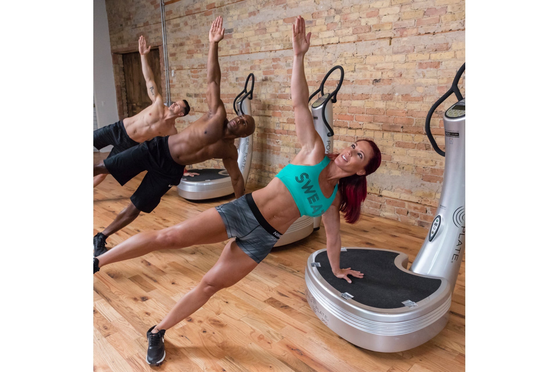 Woman in turquoise sports bra and gray shorts performing side plank on vibration plate machine in gym studio with brick wall 