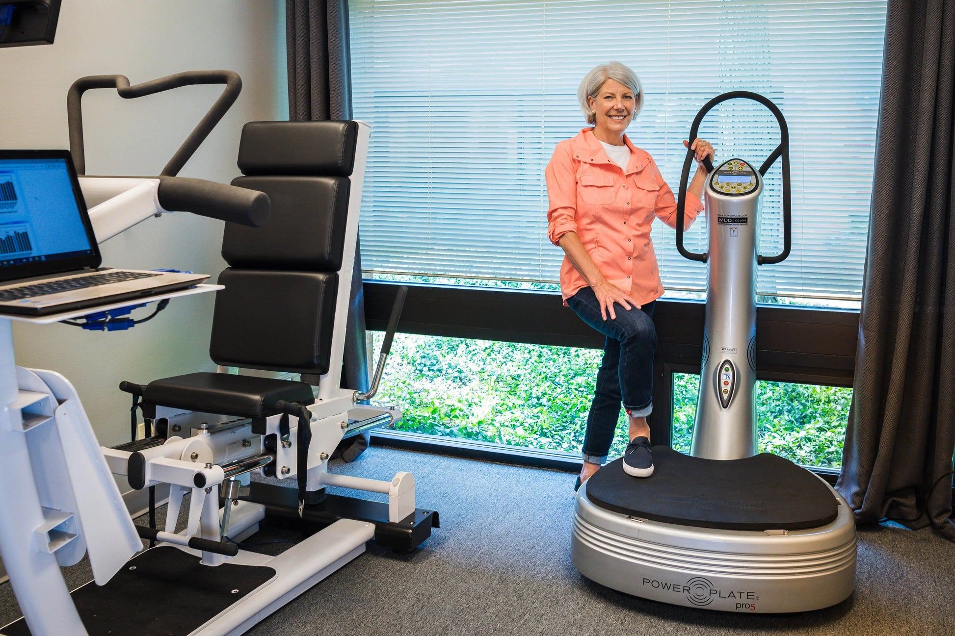 Woman in coral shirt demonstrating silver and black Power Plate Pro5 vibration plate trainer in office gym setting
