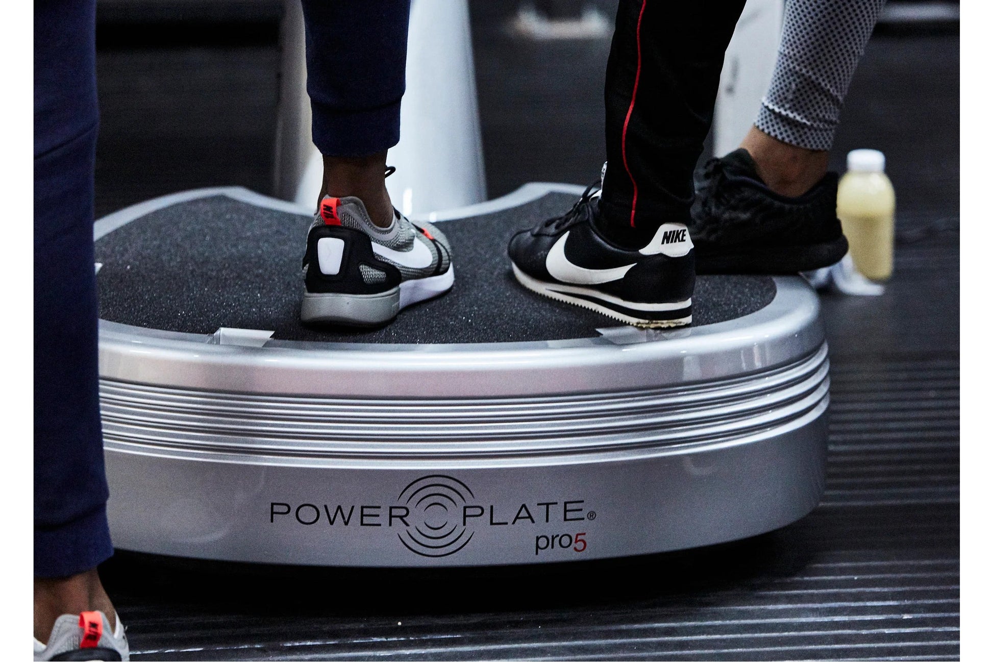 Two people wearing athletic sneakers standing on a Power Plate Pro5 vibration plate trainer with gray textured surface and wh
