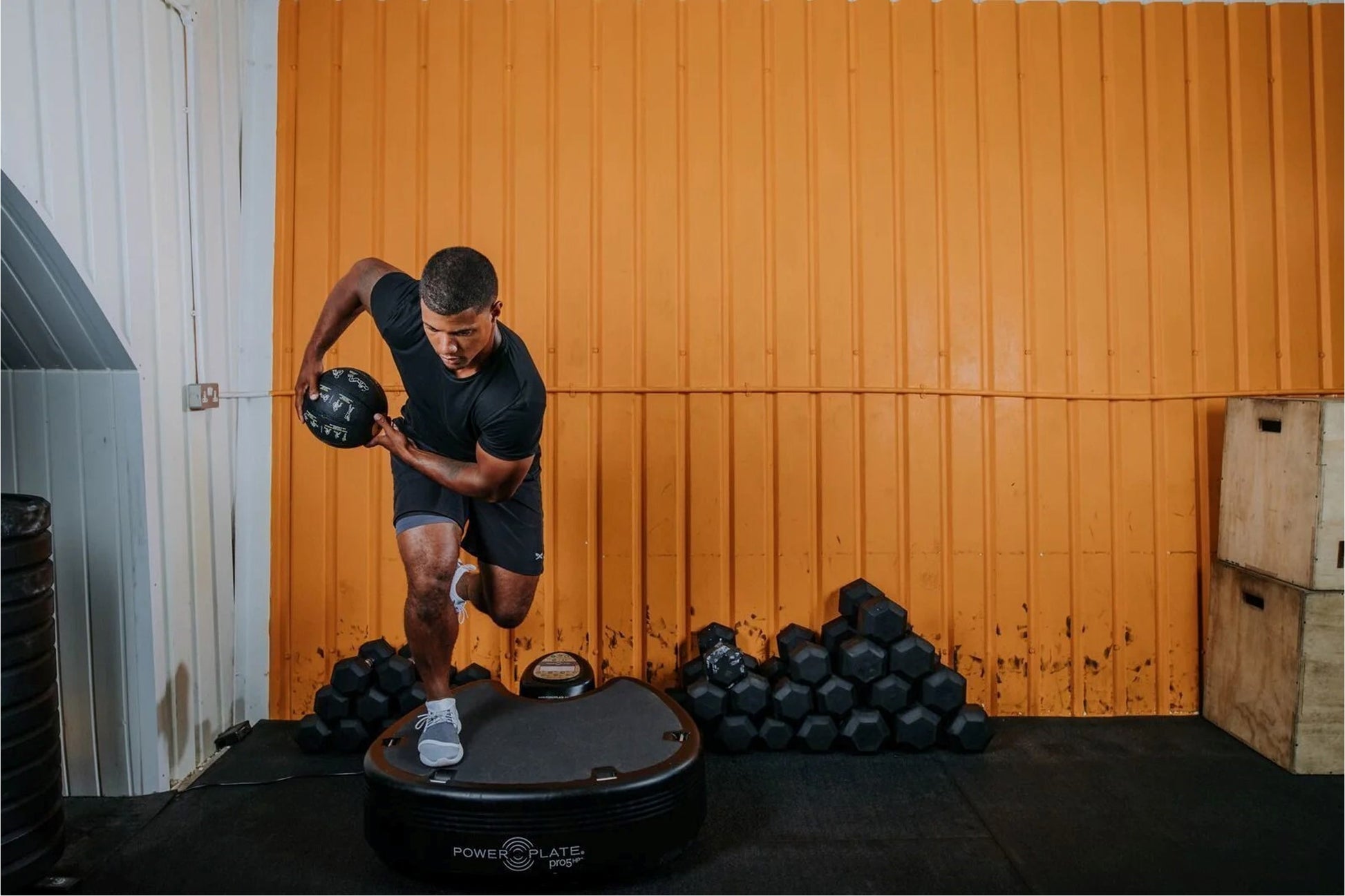 Man performing dynamic exercise on Power Plate Pro5HP vibration trainer, holding medicine ball mid-movement in gym setting wi