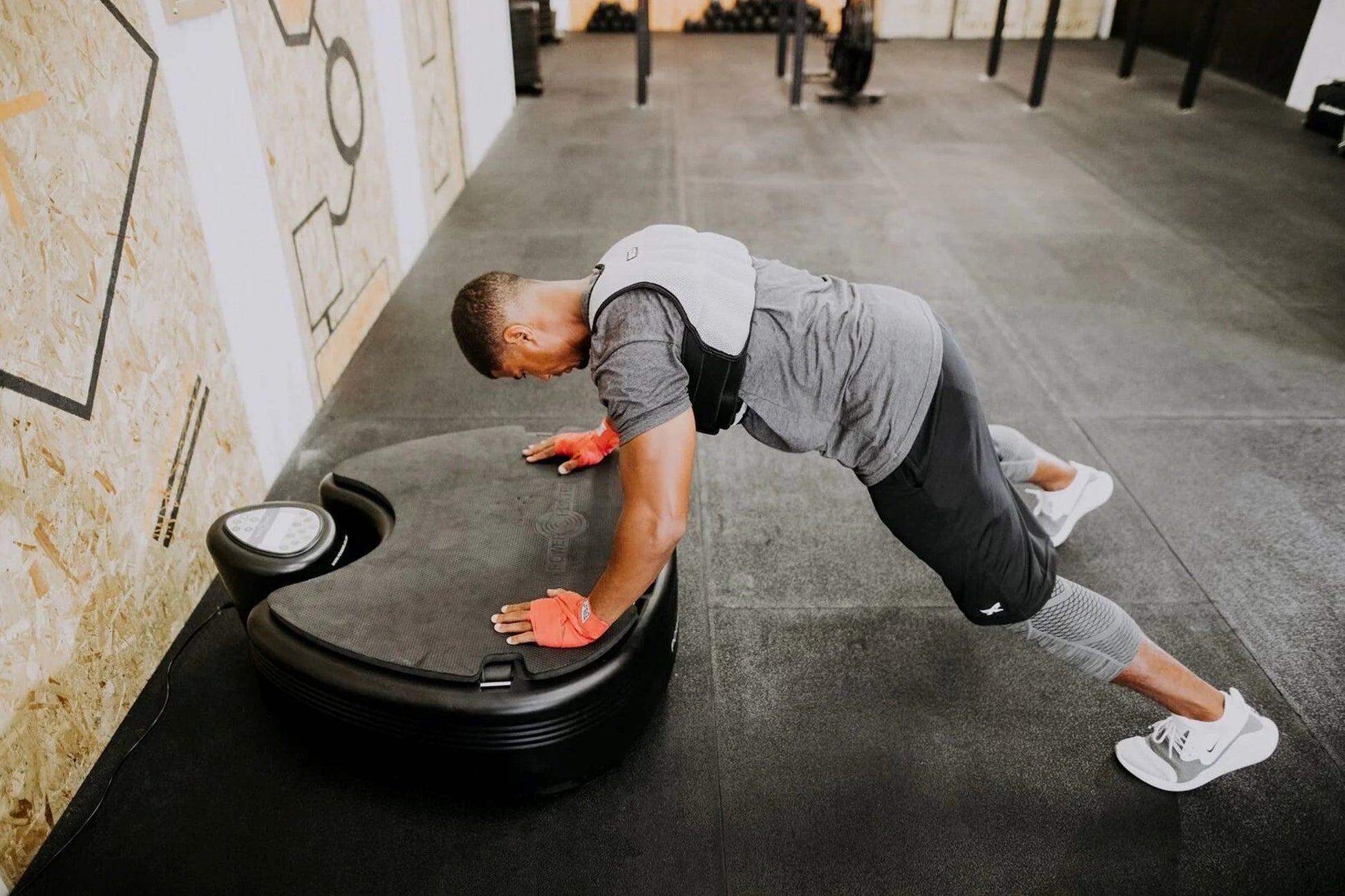 Man in white cap and gray shirt performing plank exercise on black Power Plate vibration trainer in gym setting