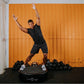 Man wearing black t-shirt and shorts performing dynamic exercise on black Power Plate vibration trainer, surrounded by dumbbe
