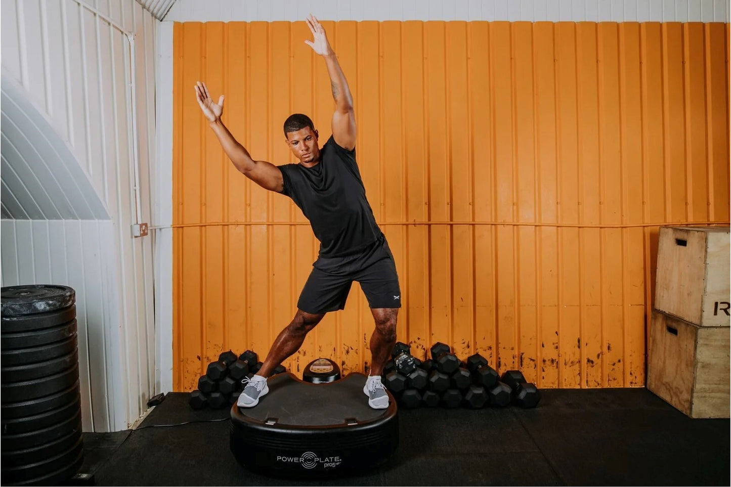 Man wearing black t-shirt and shorts performing dynamic exercise on black Power Plate vibration trainer, surrounded by dumbbe