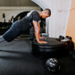 Man performing plank exercise on black Power Plate vibration trainer in gym setting, wearing black shirt and gray leggings