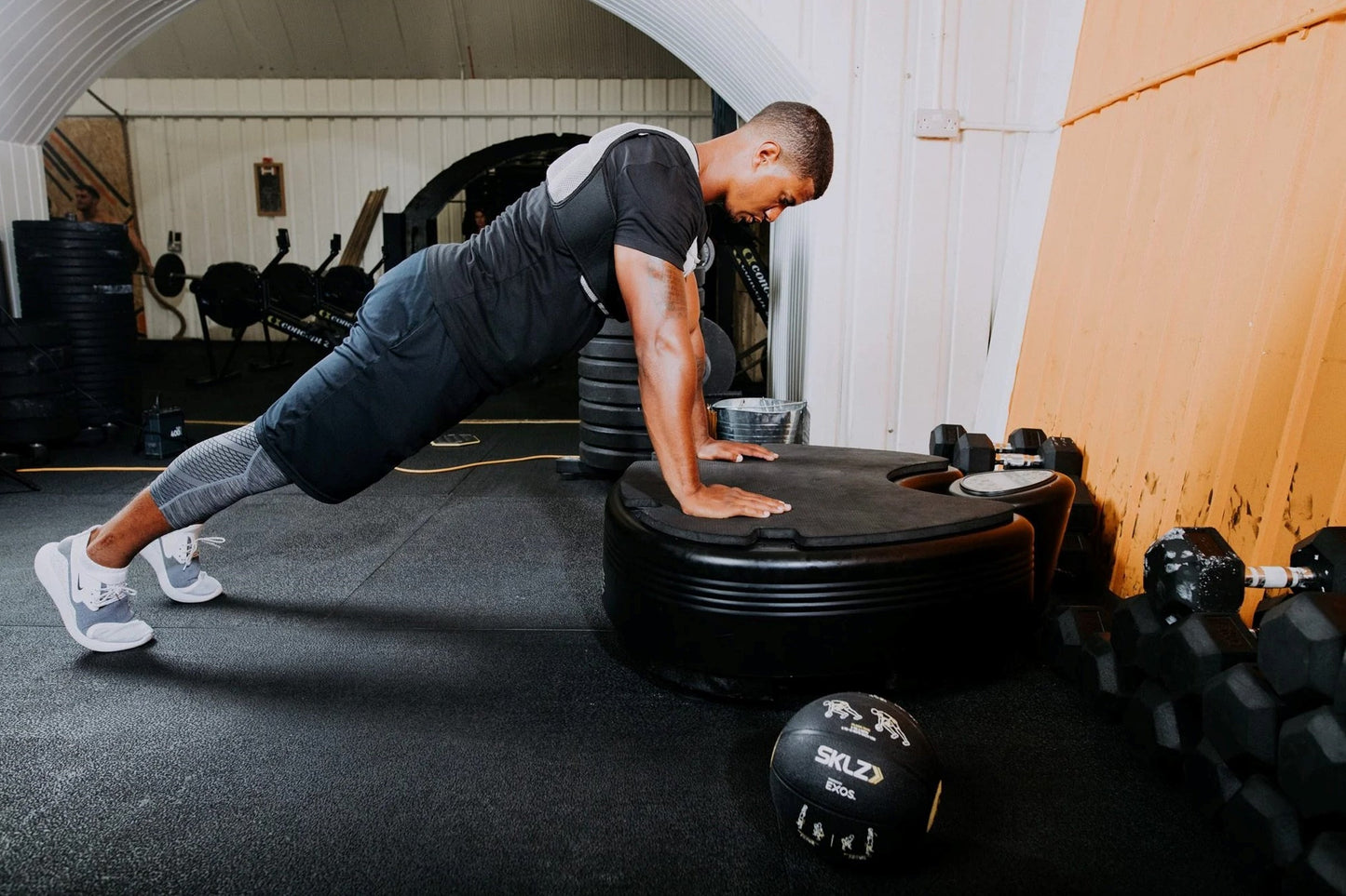 Man performing plank exercise on black Power Plate vibration trainer in gym setting, wearing black shirt and gray leggings