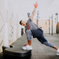 Man in gray shirt and navy shorts with coral hand wraps performing dynamic exercise move on black Power Plate device in gym s