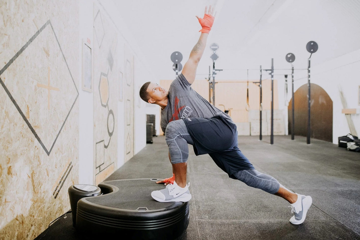 Man in gray shirt and navy shorts with coral hand wraps performing dynamic exercise move on black Power Plate device in gym s