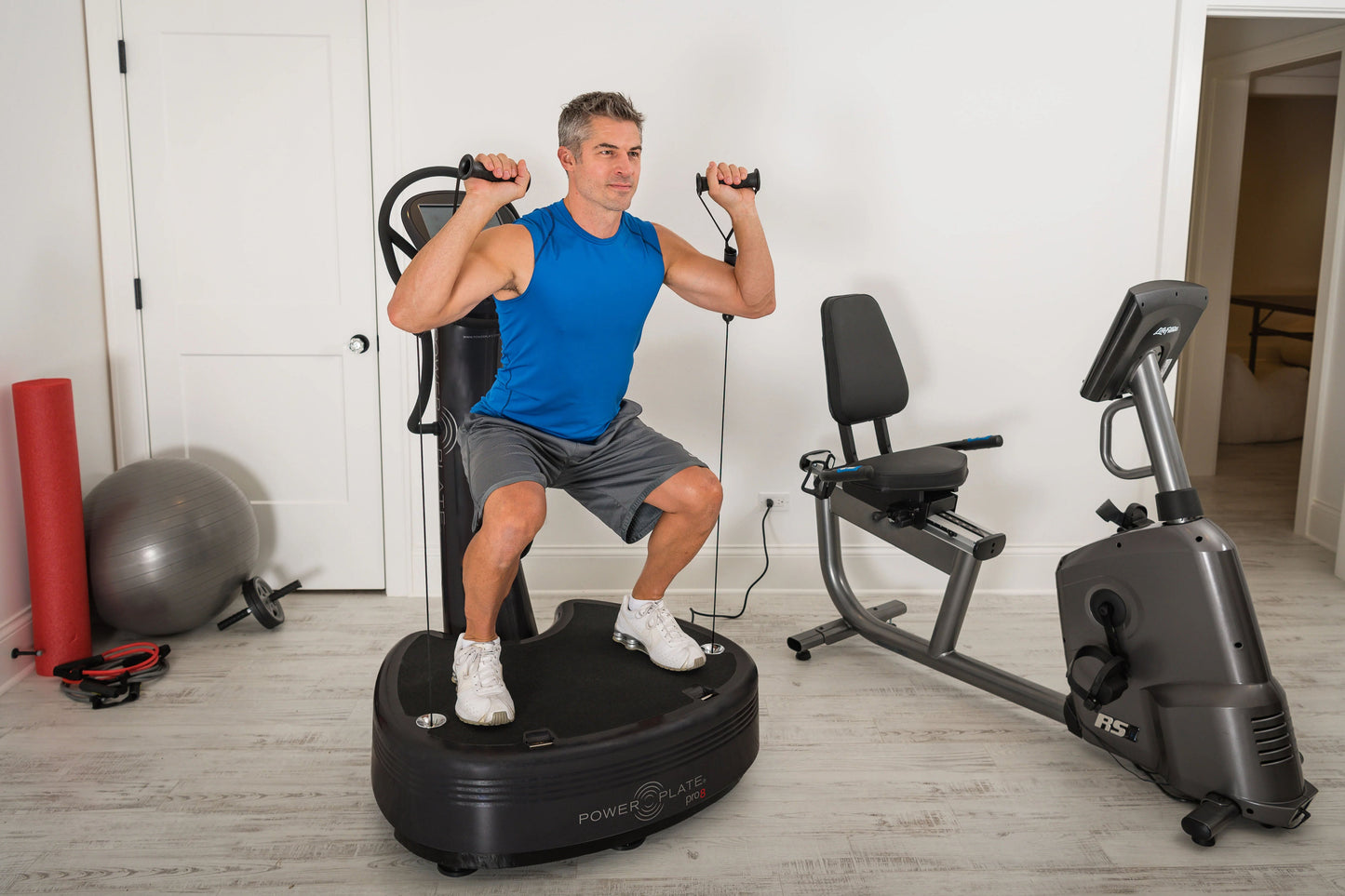 Man in blue tank top using Power Plate Pro8 vibration trainer with resistance bands in home gym setting