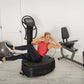 Blonde woman wearing red tank top and black leggings using black Power Plate vibration trainer in home gym setting with exerc