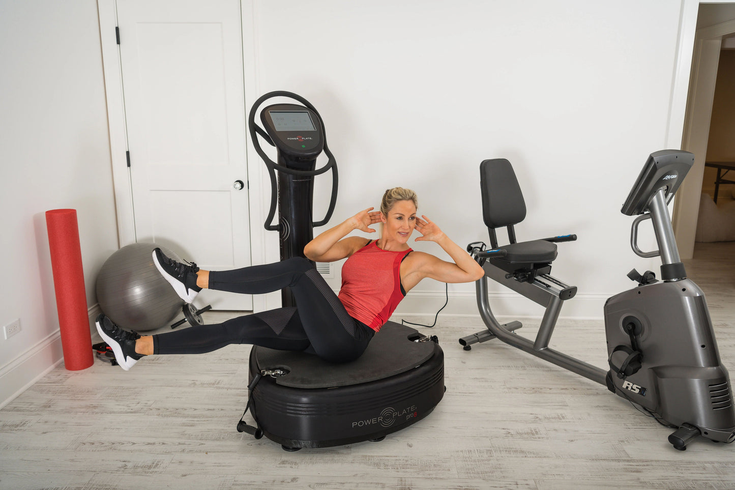Blonde woman wearing red tank top and black leggings using black Power Plate vibration trainer in home gym setting with exerc