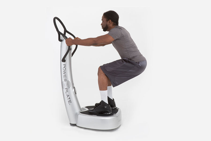 Man using white and black Power Plate vibration trainer with dual handles, wearing gray shirt and dark shorts
