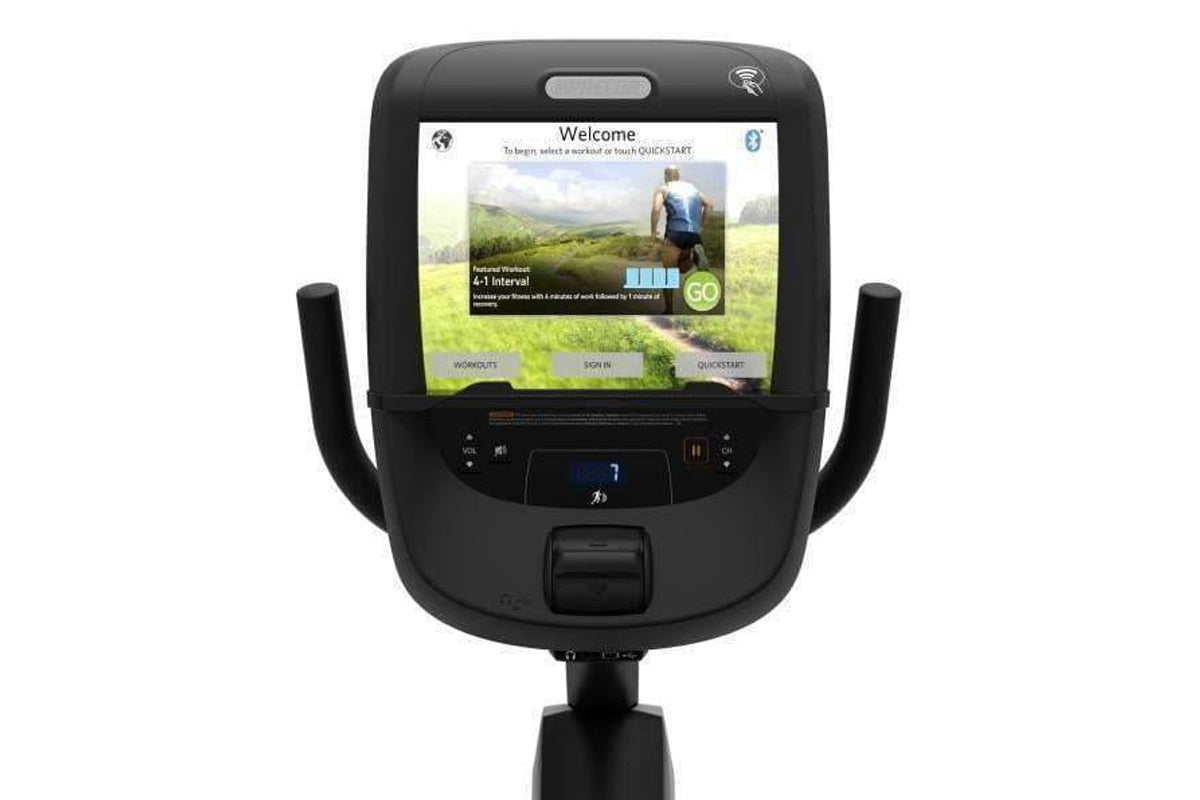 Black recumbent exercise bike with digital display screen showing welcome menu and workout options, featuring dual handles an
