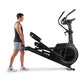 Man wearing navy tank top and gray shorts standing next to black ProForm Carbon E7 elliptical machine on white background