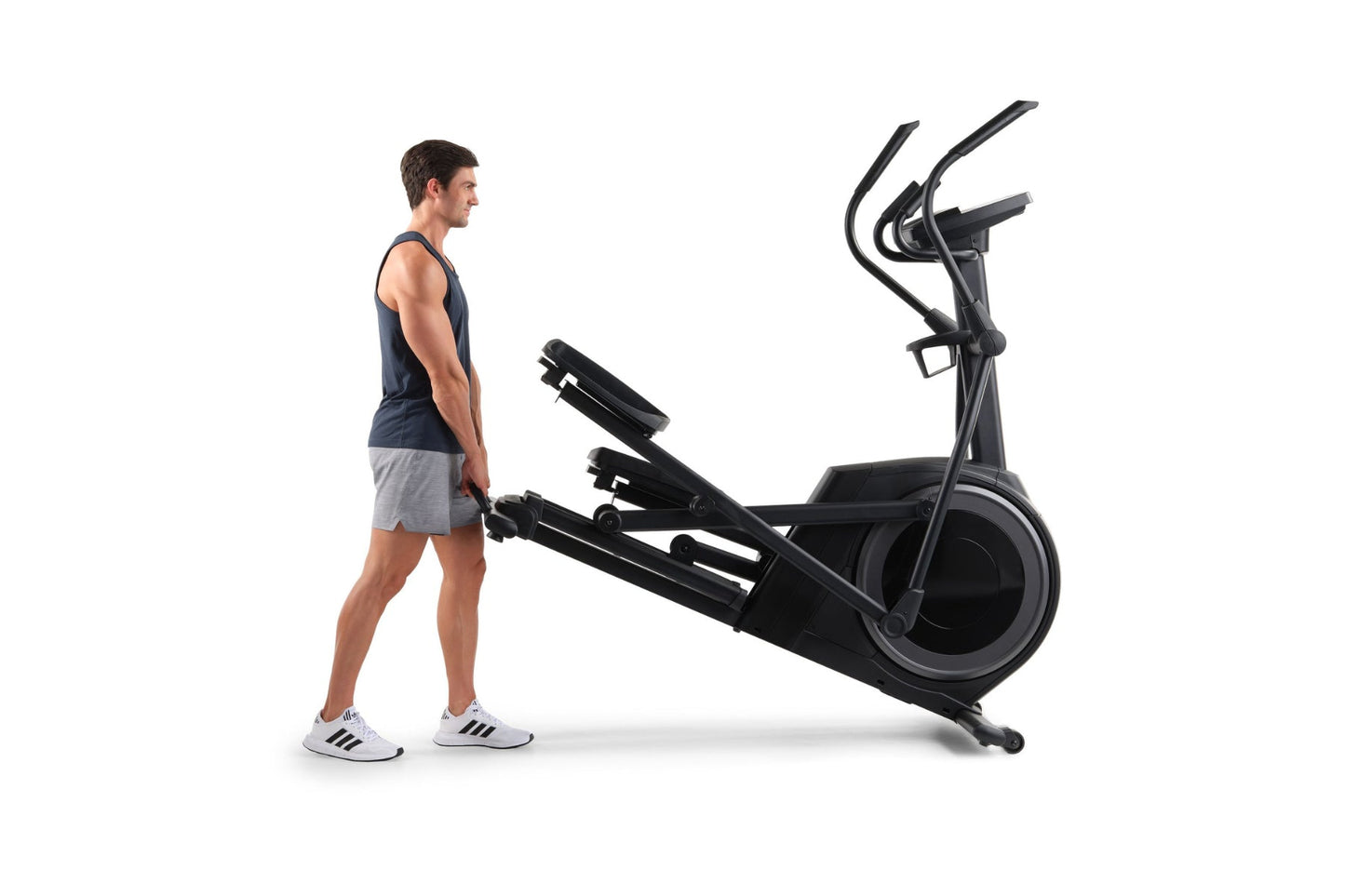 Man wearing navy tank top and gray shorts standing next to black ProForm Carbon E7 elliptical machine on white background