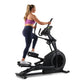 Woman in purple sports bra and navy leggings using black ProForm elliptical machine on white background