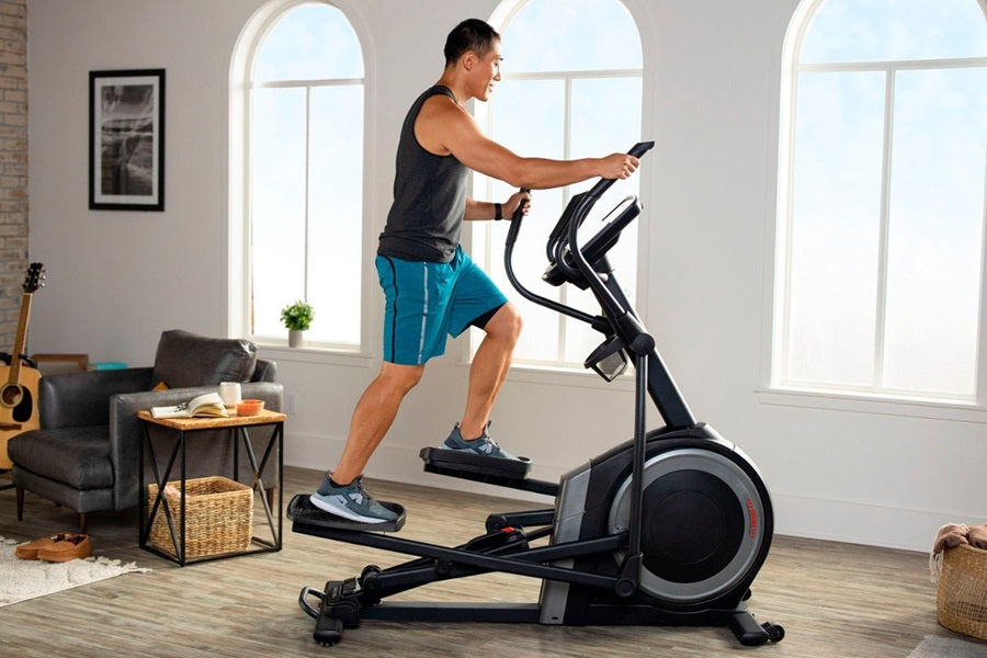 Man using black and gray elliptical machine with digital display in bright home gym with windows