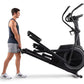 Man in navy tank top and gray shorts standing next to black Proform Plus Carbon E7 elliptical machine on white background