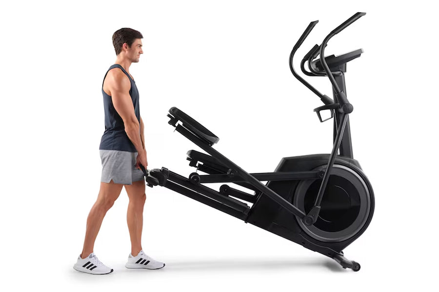 Man in navy tank top and gray shorts standing next to black Proform Plus Carbon E7 elliptical machine on white background