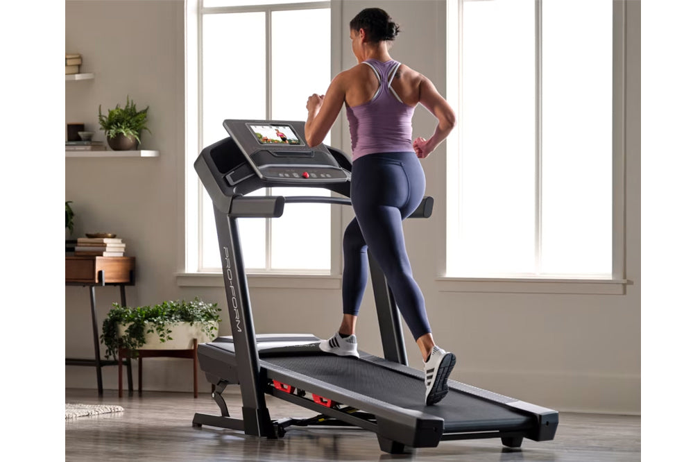 Woman wearing purple racerback tank and navy leggings running on black motorized treadmill with digital display in bright hom