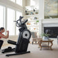 Woman in red tank top using black ProForm Pro HIIT H14 elliptical with white wheel in bright living room with fireplace