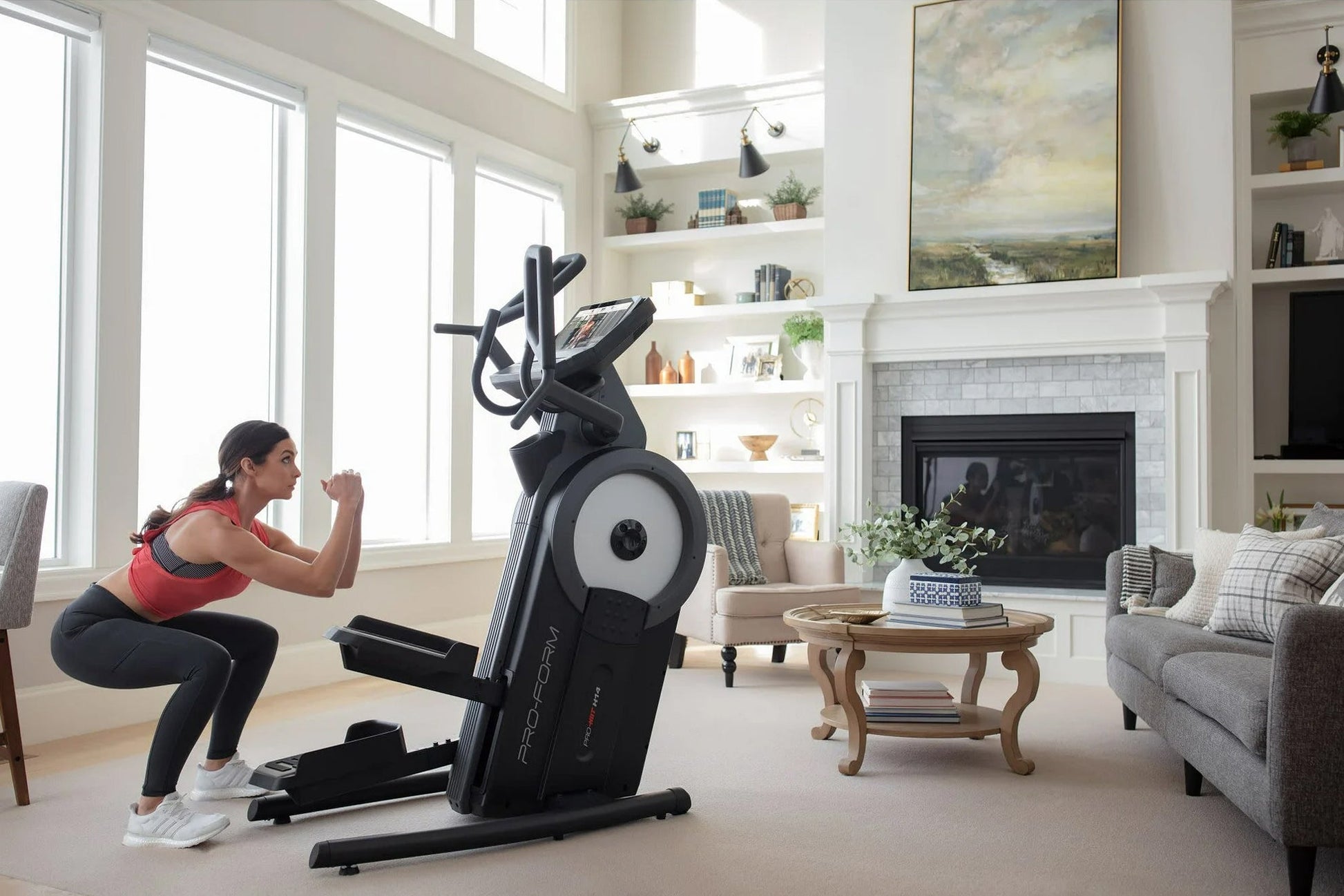 Woman in red tank top using black ProForm Pro HIIT H14 elliptical with white wheel in bright living room with fireplace