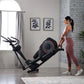 Woman in gray tank top and burgundy leggings using black ProForm Sport elliptical machine in modern home gym with brick wall 