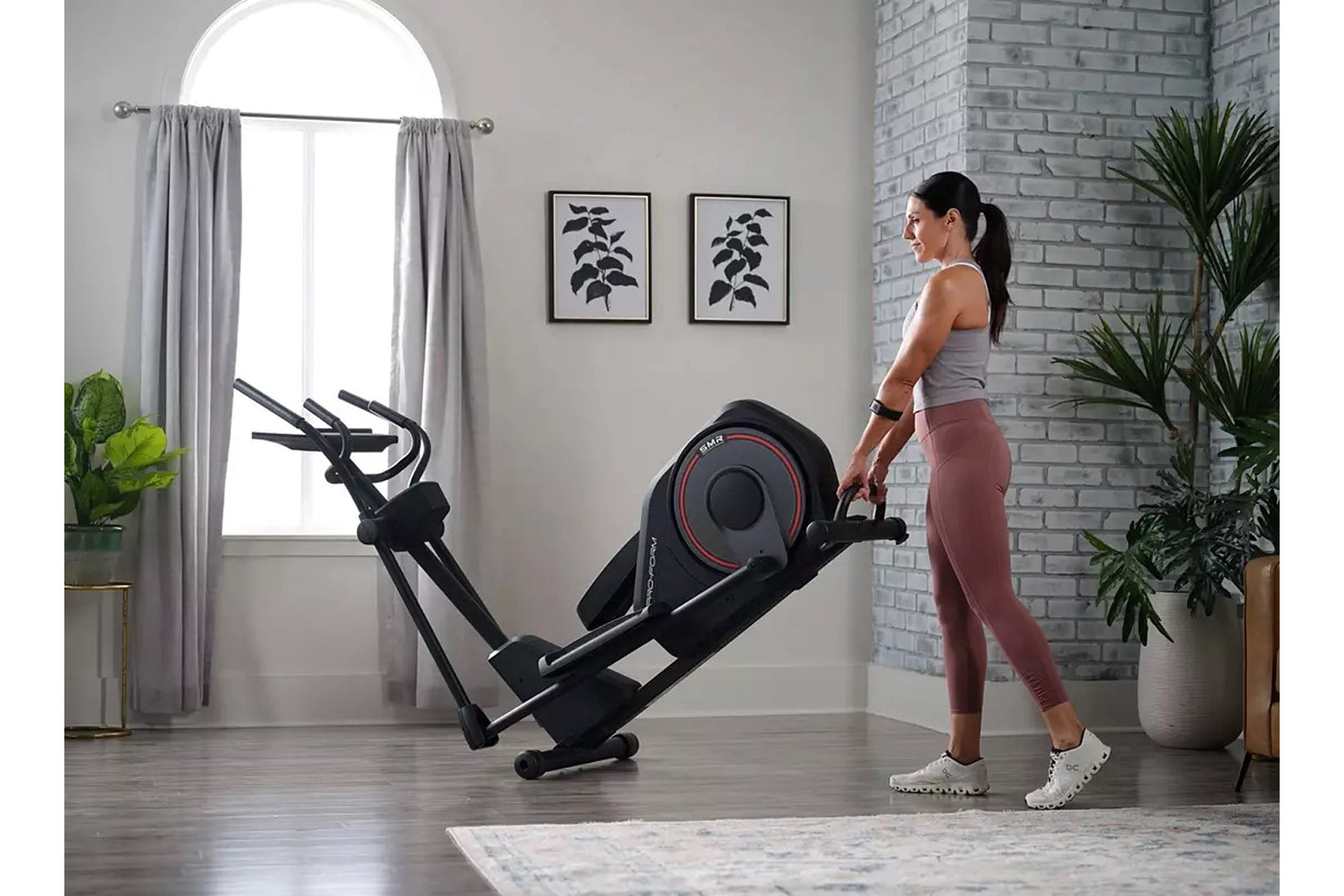 Woman in gray tank top and burgundy leggings using black ProForm Sport elliptical machine in modern home gym with brick wall 