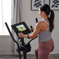 Woman wearing white tank top and burgundy leggings using black ProForm Sport Elliptical with digital display screen in home g