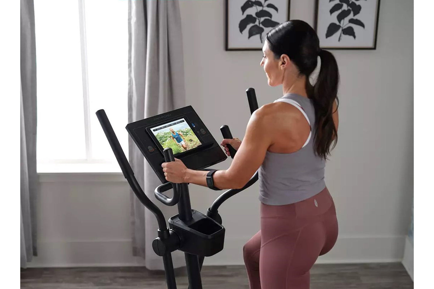 Woman wearing white tank top and burgundy leggings using black ProForm Sport Elliptical with digital display screen in home g