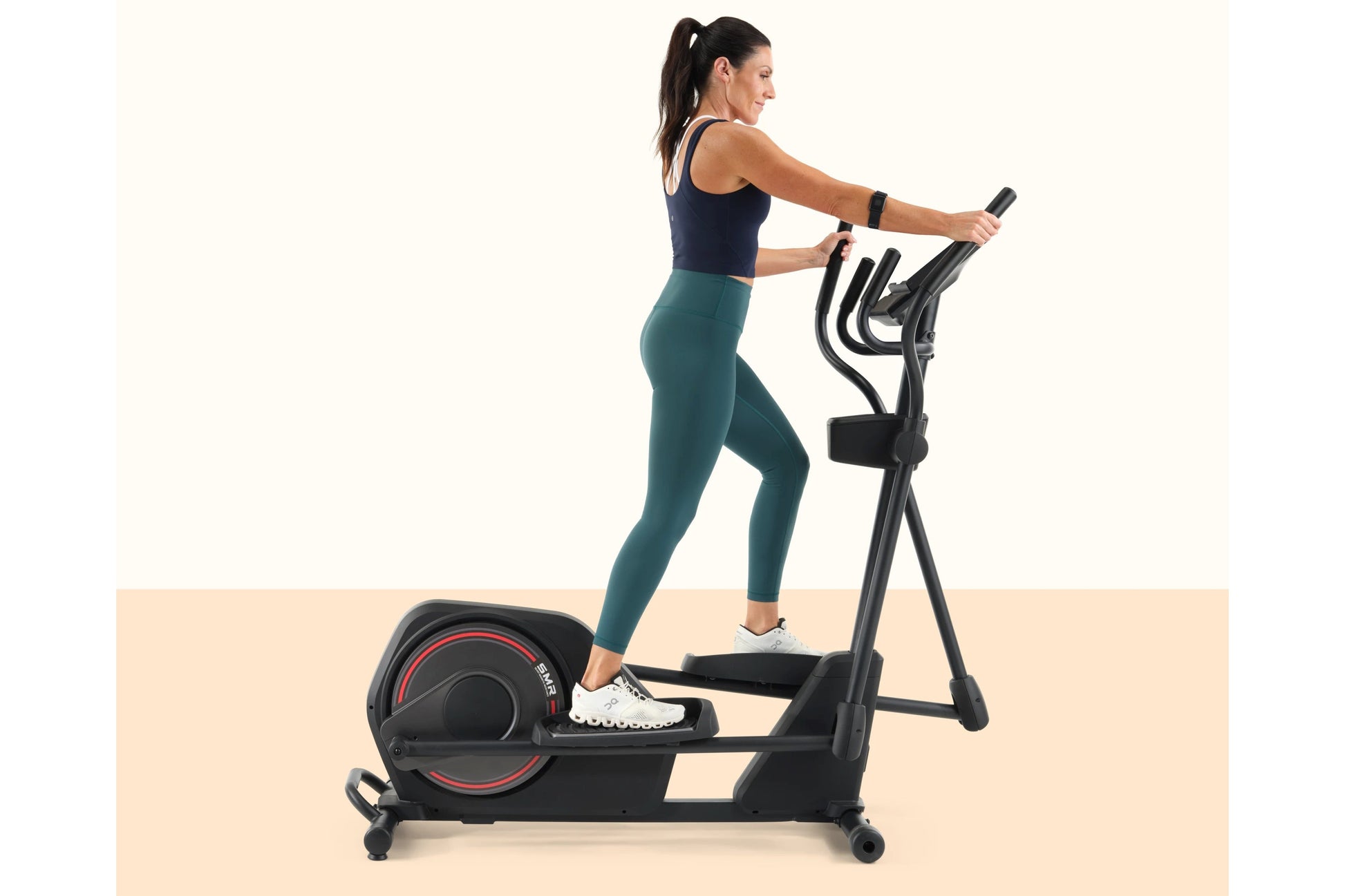 Woman using black elliptical machine with red accents, wearing navy sports bra and teal leggings, on beige background