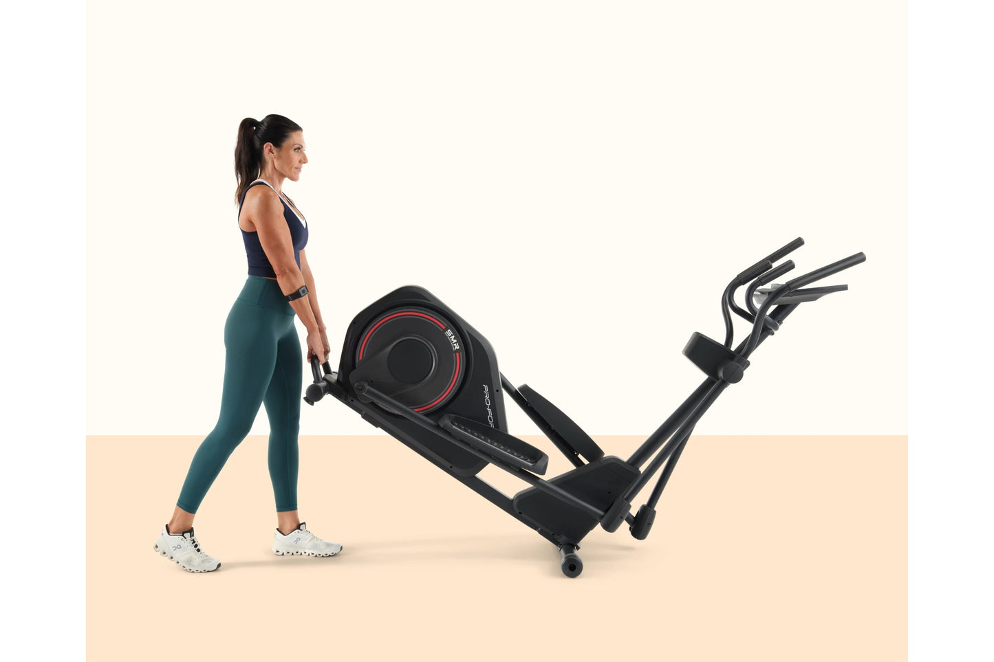 Woman wearing navy sports bra and teal leggings standing next to black ProForm elliptical machine with red accents on beige a