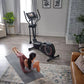 Black elliptical machine with digital display and dual handles in home gym setting, model exercising on gray mat nearby
