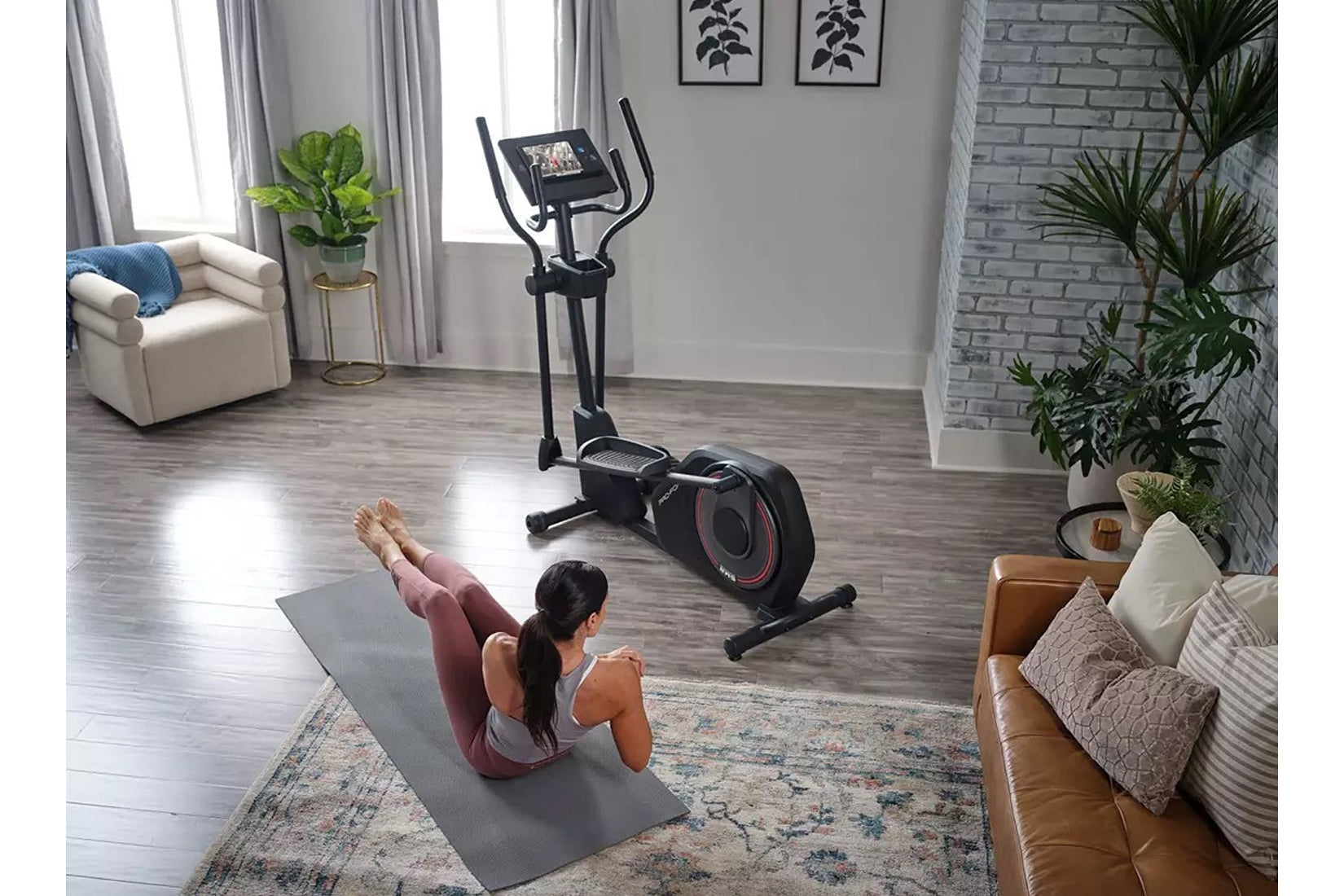 Black elliptical machine with digital display and dual handles in home gym setting, model exercising on gray mat nearby