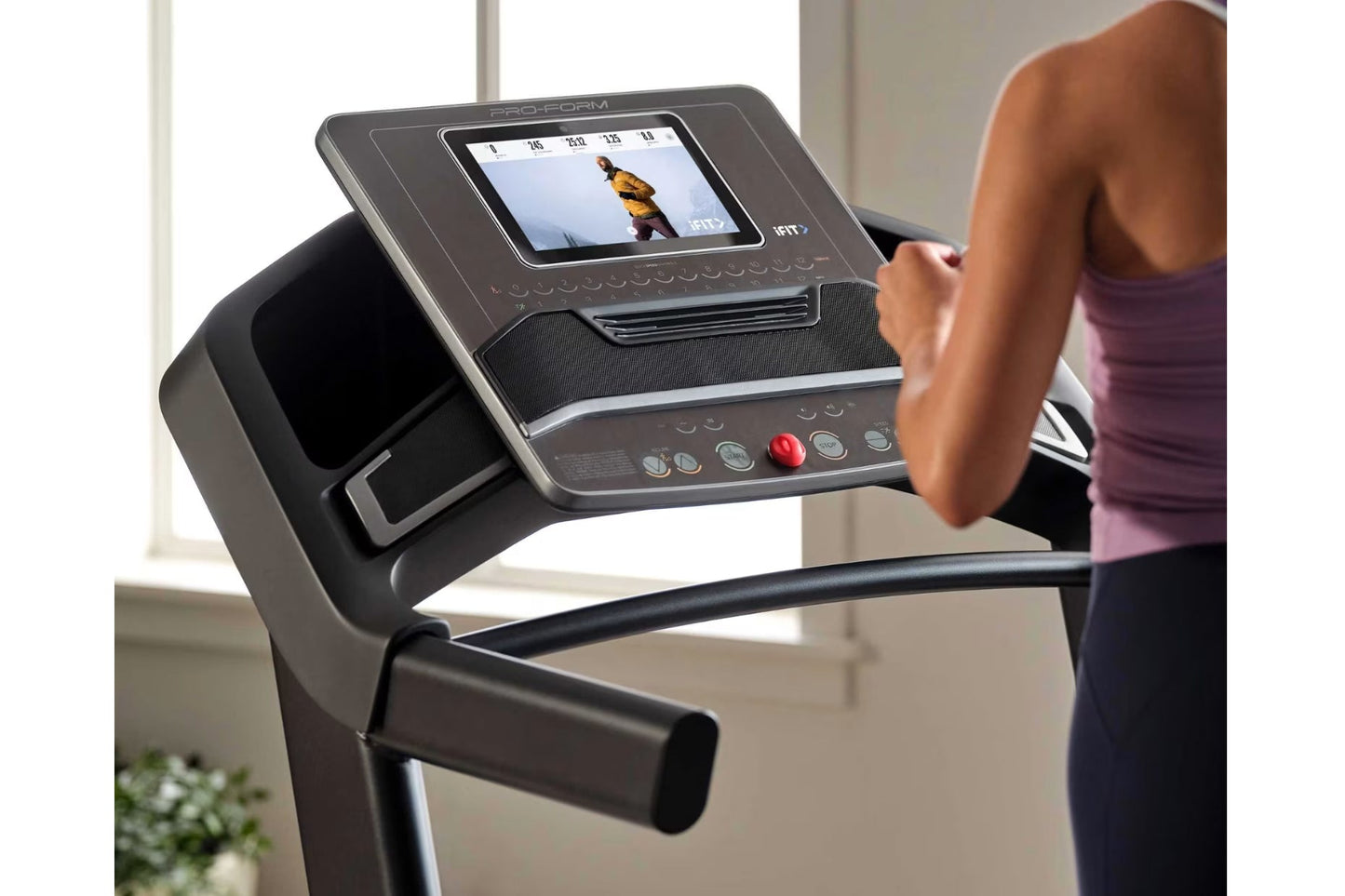 Black ProForm treadmill with digital display showing workout video, control buttons, and red emergency stop button, woman usi
