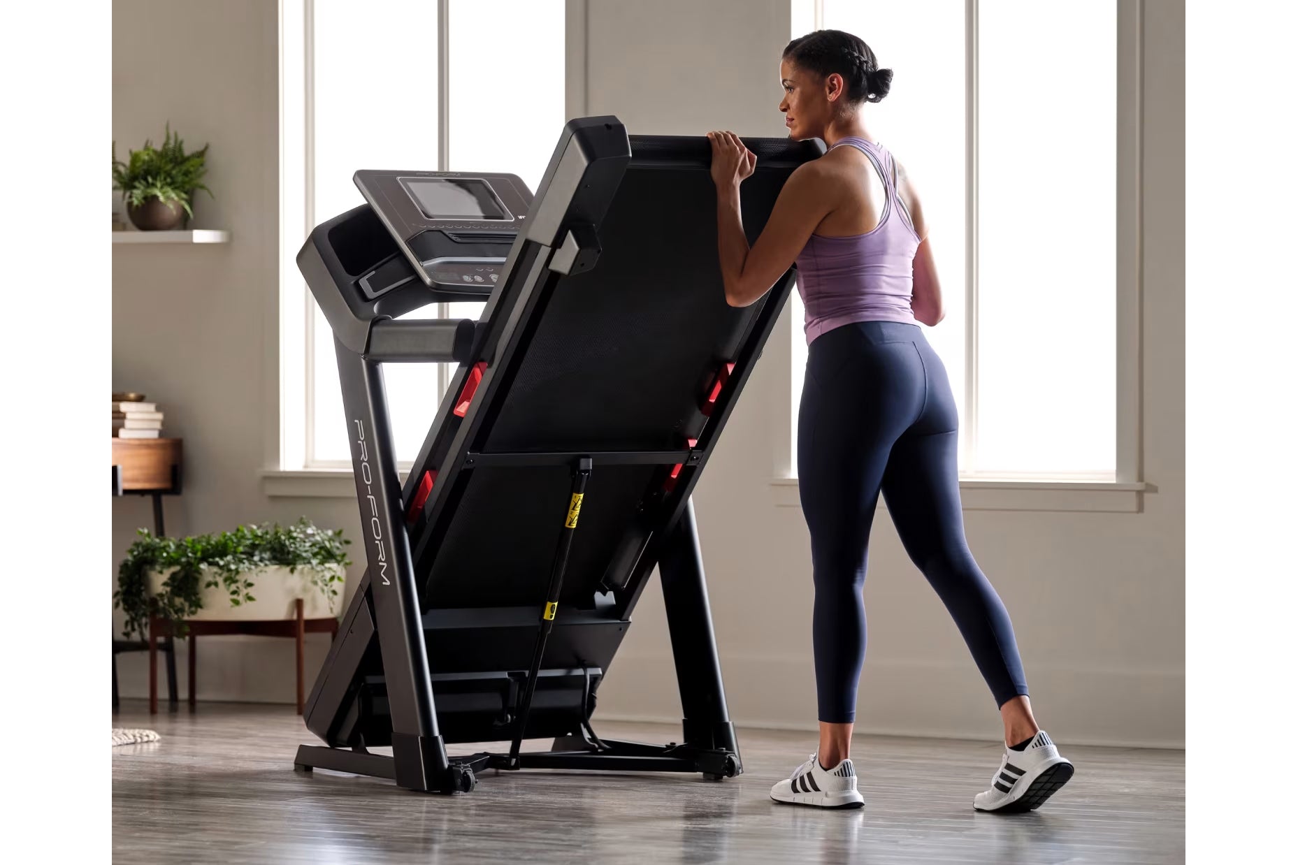 Woman in purple tank top and navy leggings running on black ProForm treadmill with digital display in bright home gym