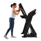 Woman using black ProForm Trainer 1000 treadmill with digital display, wearing white tank top and navy leggings