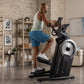Man in white tank top and blue shorts using black ProForm elliptical machine with silver flywheel in home gym with brick wall