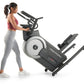 Woman wearing burgundy sports bra and gray leggings using black and gray ProForm elliptical machine with white wheel accent o