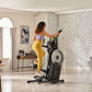 Woman in lavender sports bra and yellow leggings using black ProForm Trainer HL HIIT elliptical in modern living room