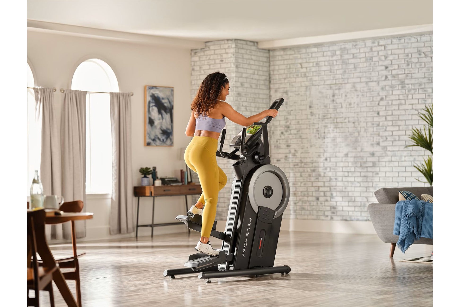Woman in lavender sports bra and yellow leggings using black ProForm Trainer HL HIIT elliptical in modern living room
