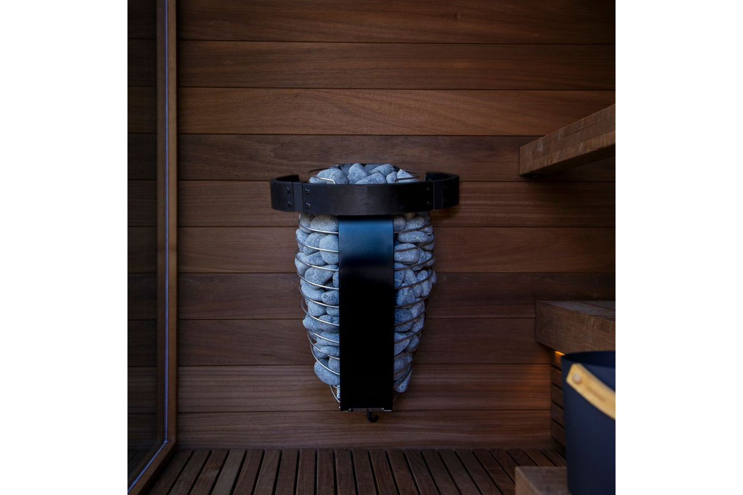 Black metal sauna heater bucket with gray stones, mounted on wooden wall inside sauna