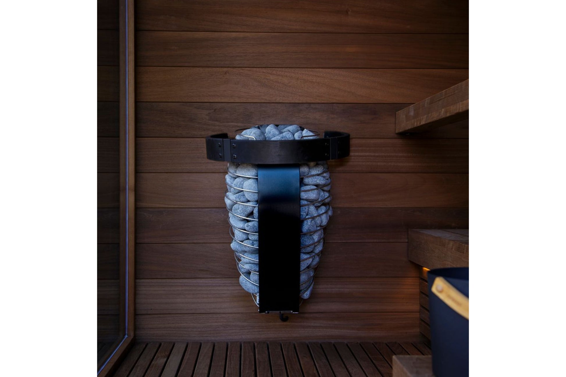 Black metal sauna heater bucket with gray stones, mounted on wooden wall inside sauna