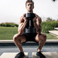 Man holding black NÜOBELL adjustable dumbbell during outdoor squat exercise by pool with landscape background