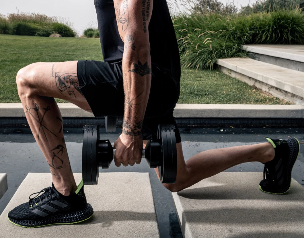 Person performing single-leg dumbbell exercise wearing black shorts and black sneakers with neon trim, holding adjustable bla
