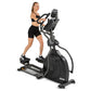 Woman in black sports bra and shorts using black and silver Spirit elliptical machine with digital display on white backgroun