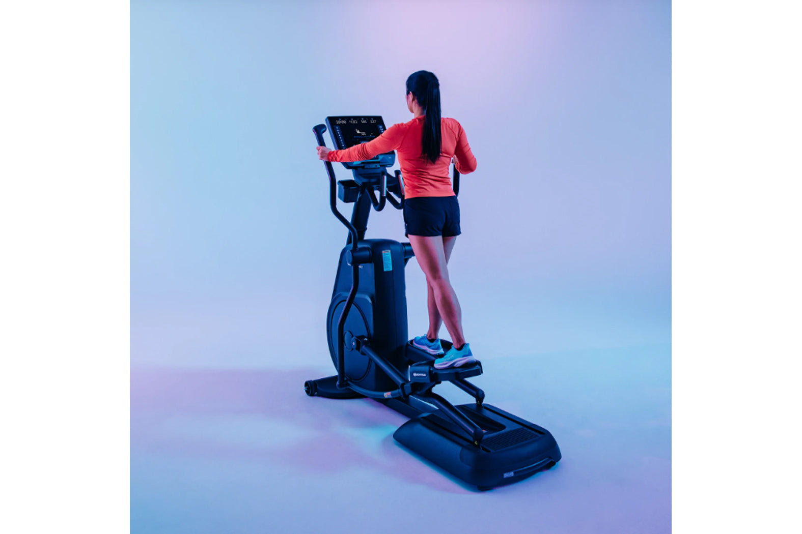 Woman wearing coral long-sleeve top and navy shorts using blue elliptical trainer with digital display against gradient blue 