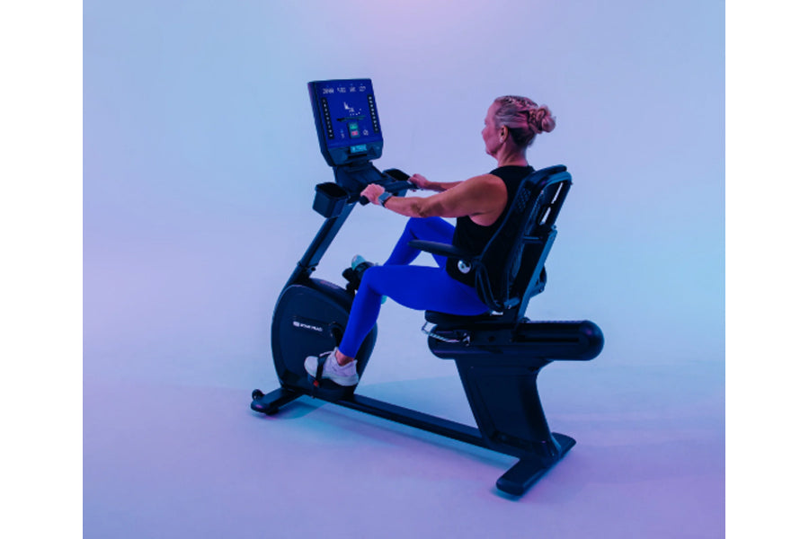 Woman using black Star Trac recumbent bike with digital display console on gradient blue and purple background